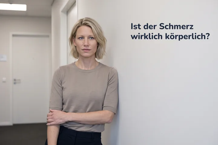 Ist der Schmerz wirklich körperlich? — eine Frau hält im Bürogang inne, den Unterarm quer über dem Bauch